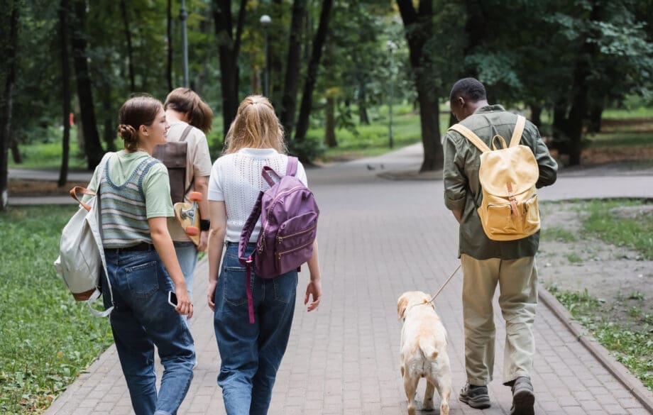A group of college-age students walking through a campus park with one student leading a dog on a leash, illustrating life at colleges that allow pets in dorms.